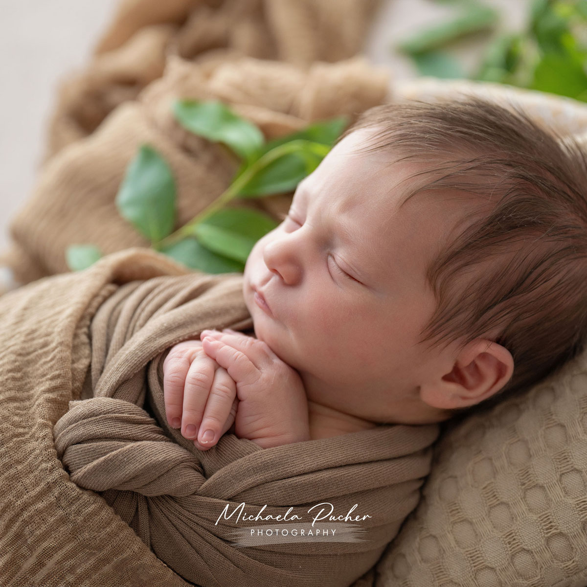 Nahaufnahme eines schlafenden Neugeborenen, eingewickelt in braune Tücher, die Hände sanft aneinandergelegt, mit grünen Blättern im Hintergrund.